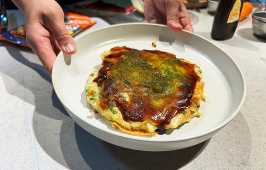 Person holding a plate with an okonomiyaki Japanese pancake on top, covered with sauce and aonori