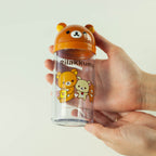 Hand holding a clear bottle with Rilakkuma design on an orange lid against a white background