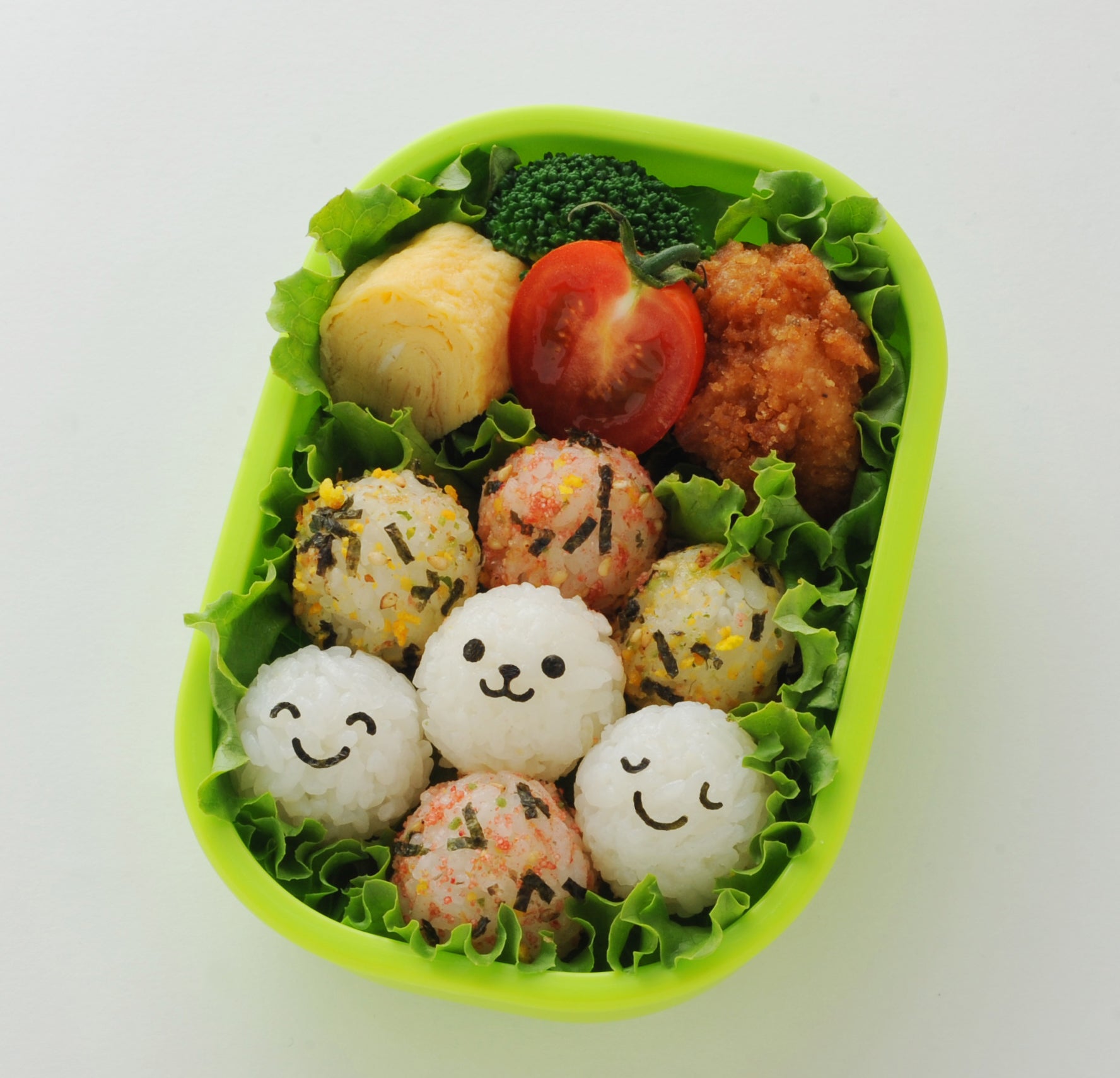Overhead view of a green oval bento box filled with plain and furikake-seasoned baby onigiri, three with cute nori smiley and bear faces, alongside tamagoyaki, cherry tomato, fried chicken, and broccoli on lettuce