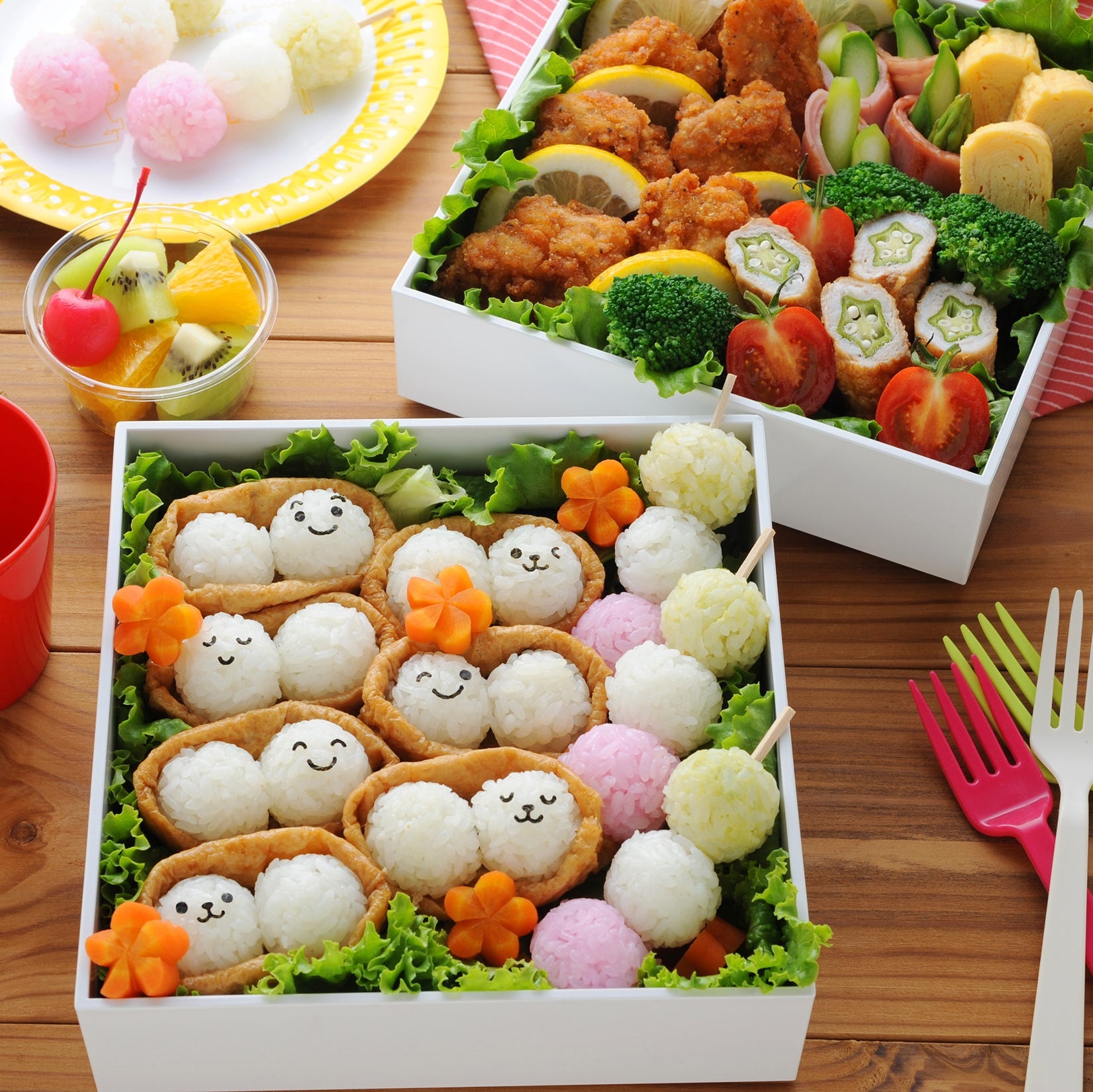 Open white square bento box filled with cute face-stamped baby onigiri nestled in aburaage pouches with carrot flowers, colorful rice balls on skewers, and a second bento box with fried chicken, broccoli, and rolled vegetables in the background
