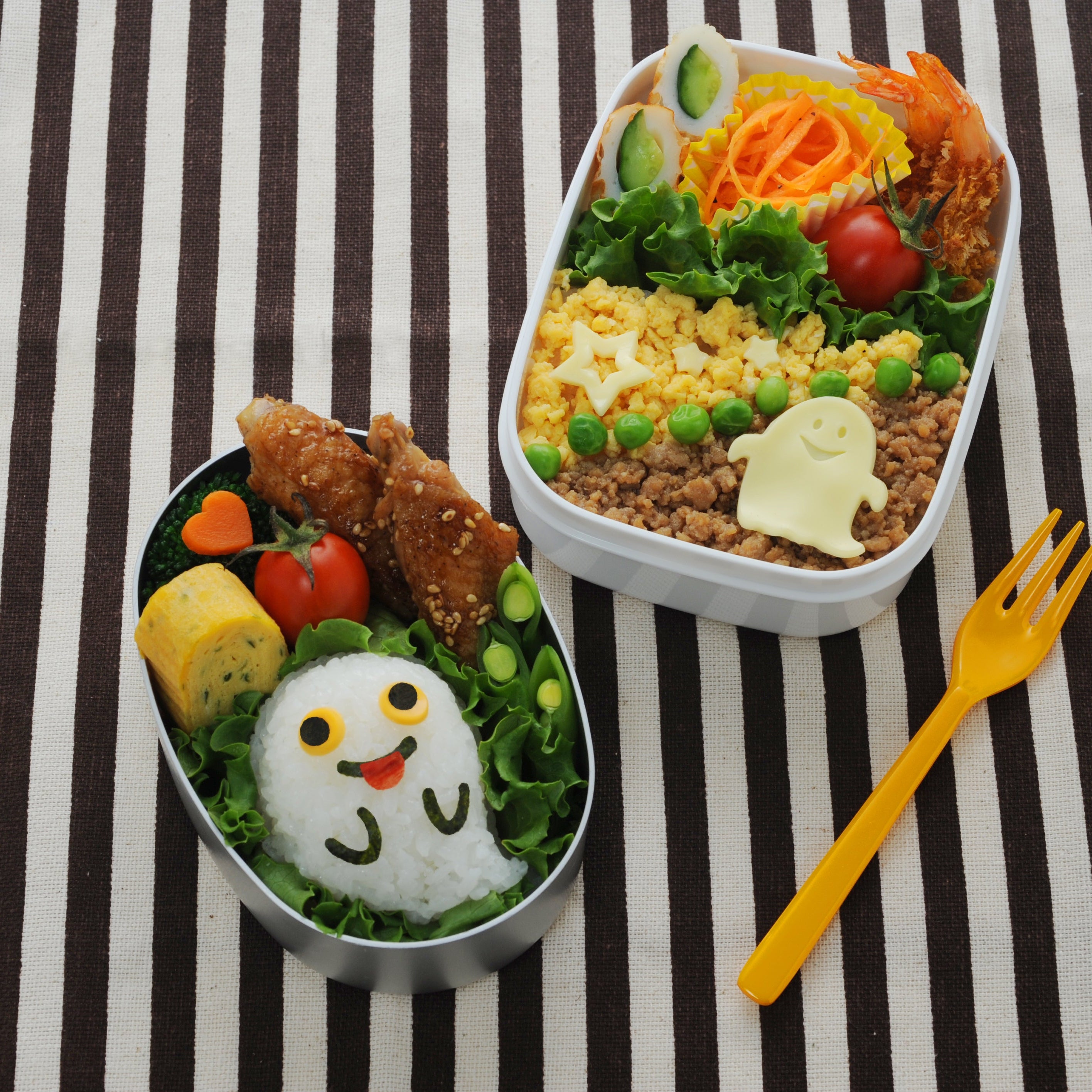 Round silver bento box with a large ghost-face onigiri, tamagoyaki, fried chicken, cherry tomatoes, and heart-shaped carrots, beside a white rectangular bento box with a ghost-shaped cheese decoration on soboro rice, peas, cucumber rolls, and shrimp, on a black and white striped cloth