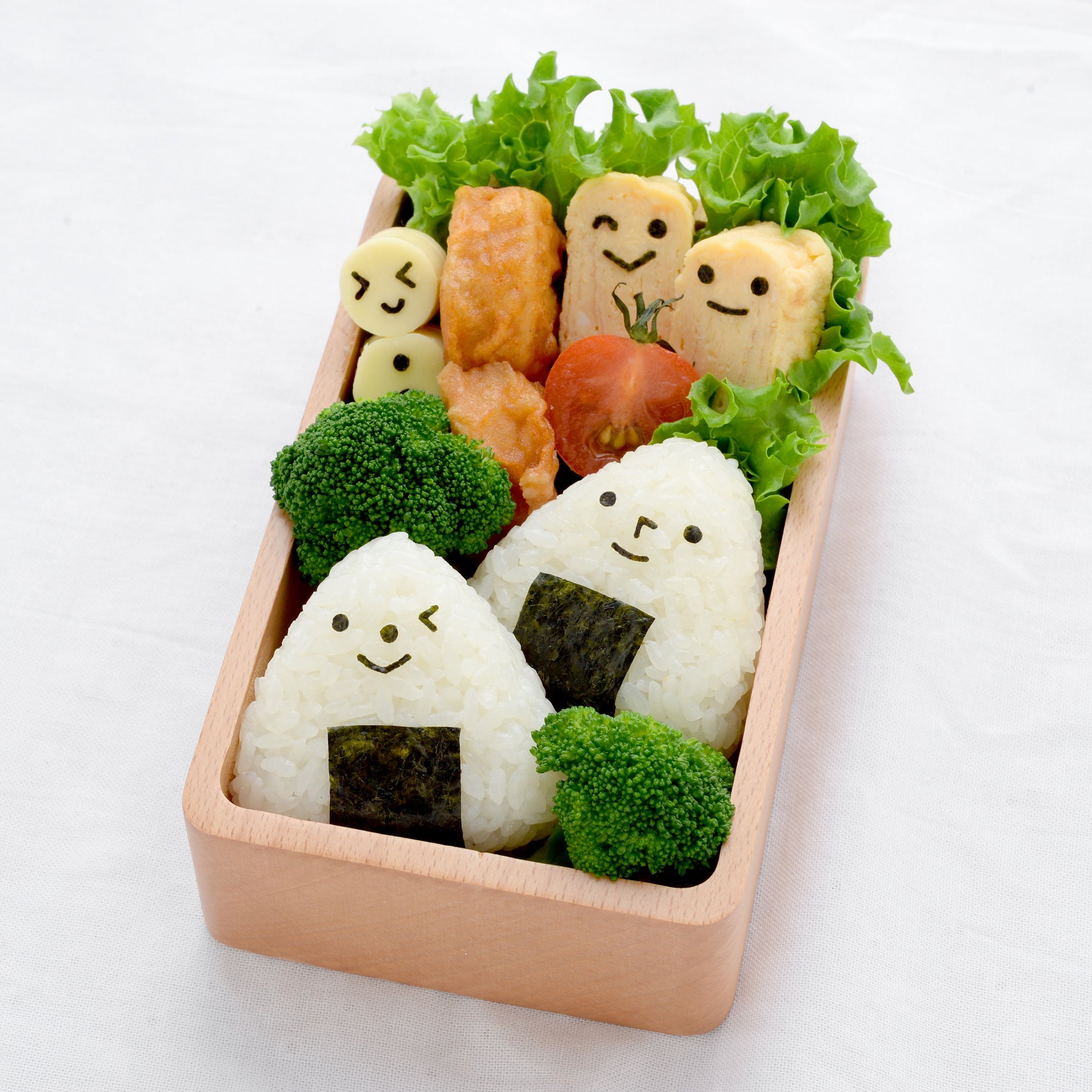 Wooden magewappa bento box filled with two triangle onigiri decorated with cute nori smiley faces using the Nico Nico Nori Puncher, alongside broccoli, cherry tomato, tamagoyaki, and face-stamped cheese and fish cake pieces, on a white cloth
