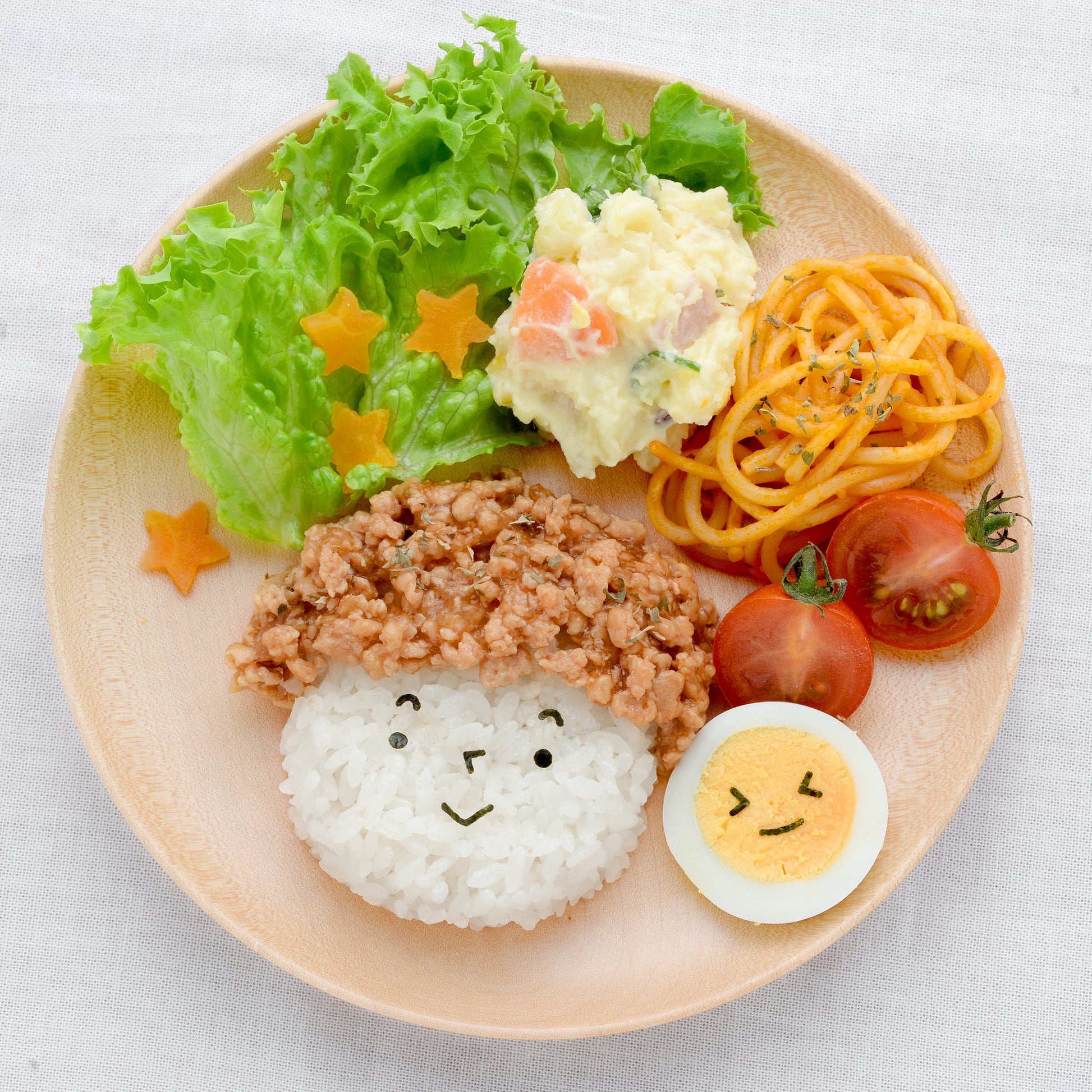 Children's lunch plate with a large round rice ball topped with soboro and a cute nori smiley face, a halved boiled egg with a nori winking face, cherry tomatoes, potato salad, spaghetti, lettuce, and star-shaped carrots, on a beige plate
