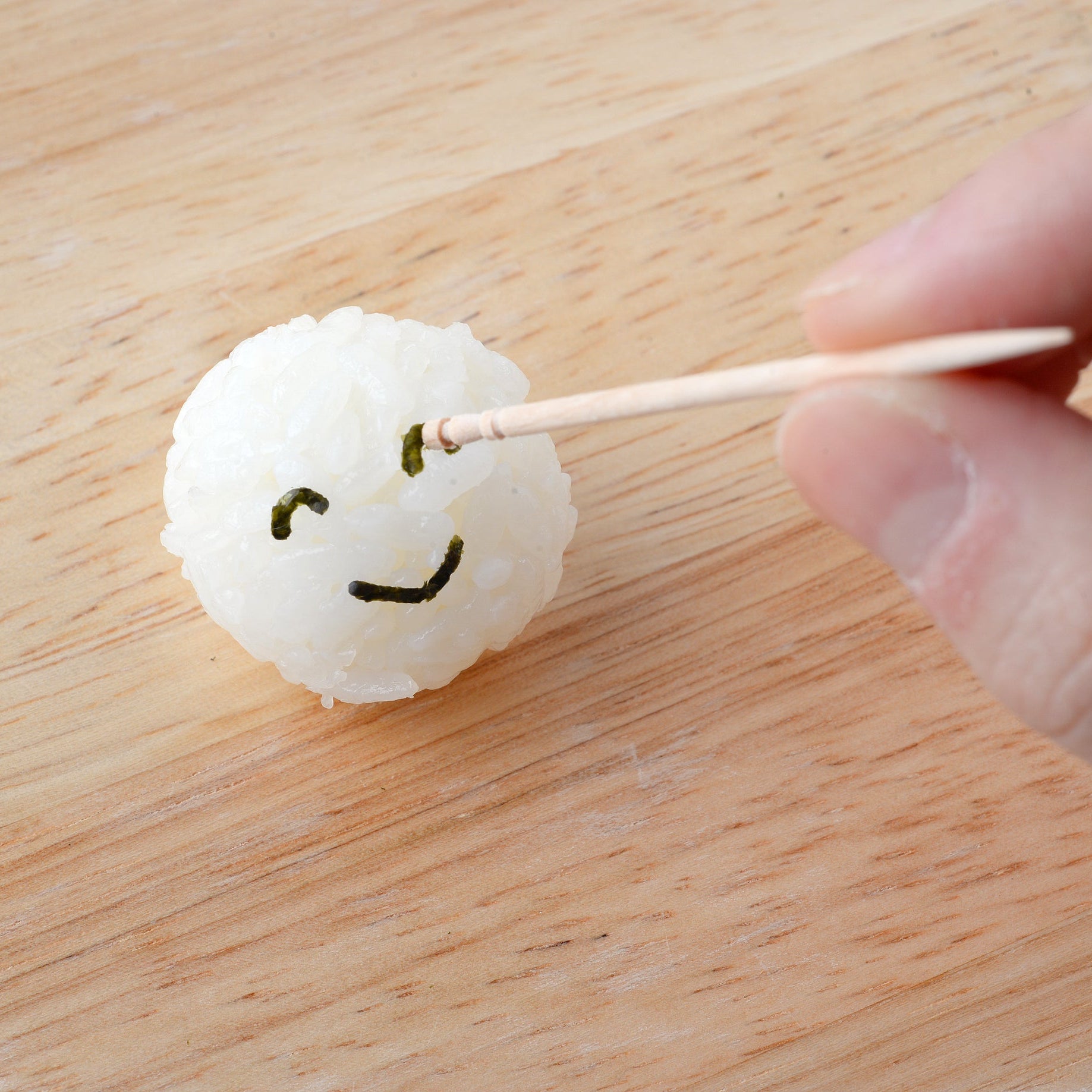 Hand using a toothpick to carefully place a small nori winking-face cutout onto a round white rice ball on a wooden surface, showing the Nico Nico Nori Puncher Mini result

