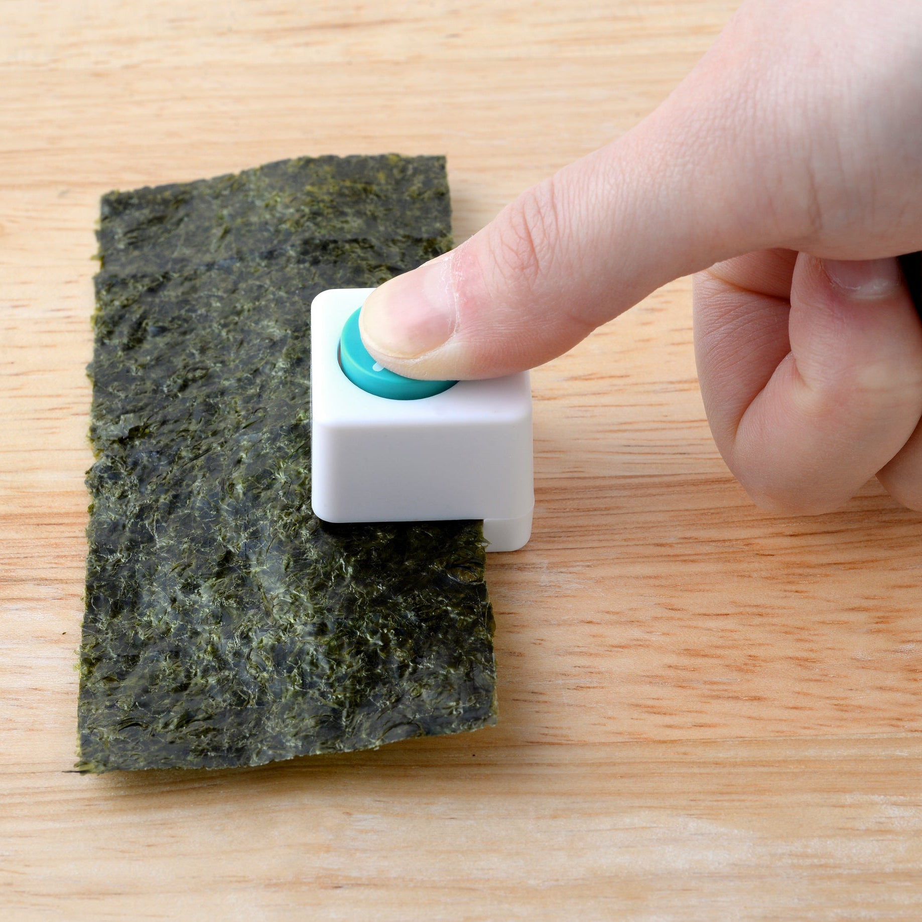 Hand pressing the teal Nico Nico Nori Puncher Mini onto a flat sheet of nori seaweed on a wooden cutting board, demonstrating how to punch out a cute face shape
