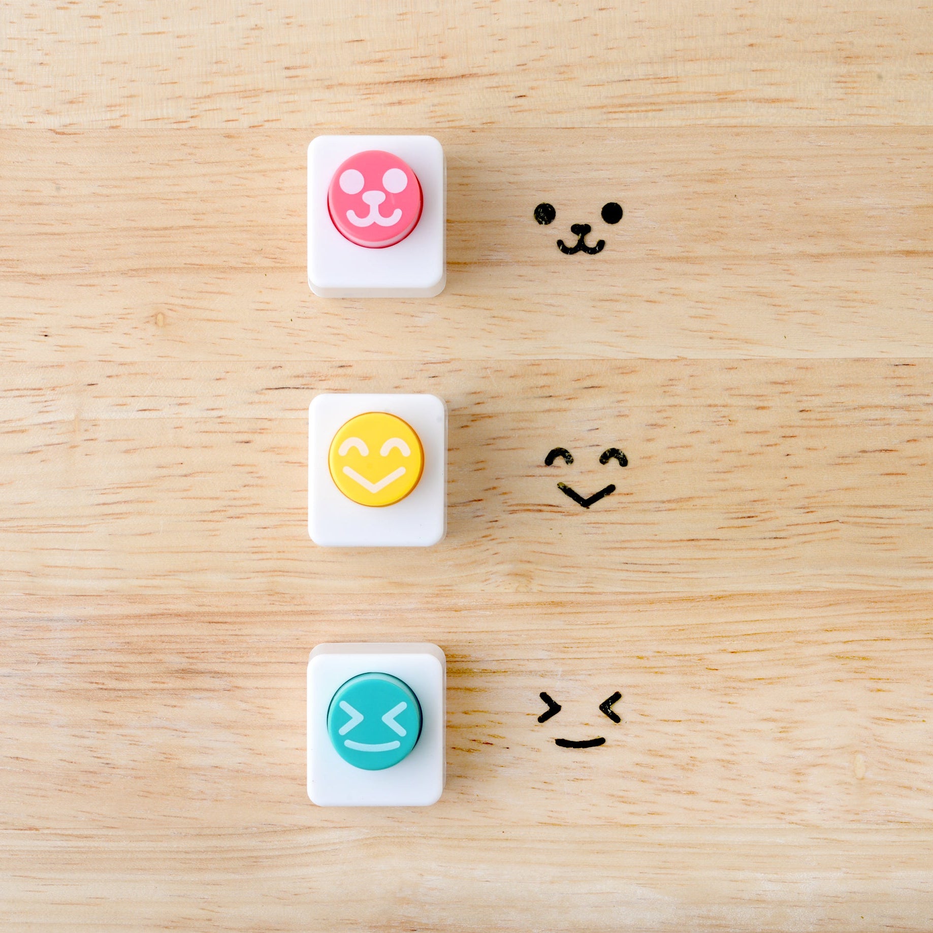 Three Arnest Nico Nico Nori Puncher Mini tools on a wooden surface – pink bear-face punch, yellow happy-face punch, and teal winking-face punch – each shown beside their corresponding nori cutout impression
