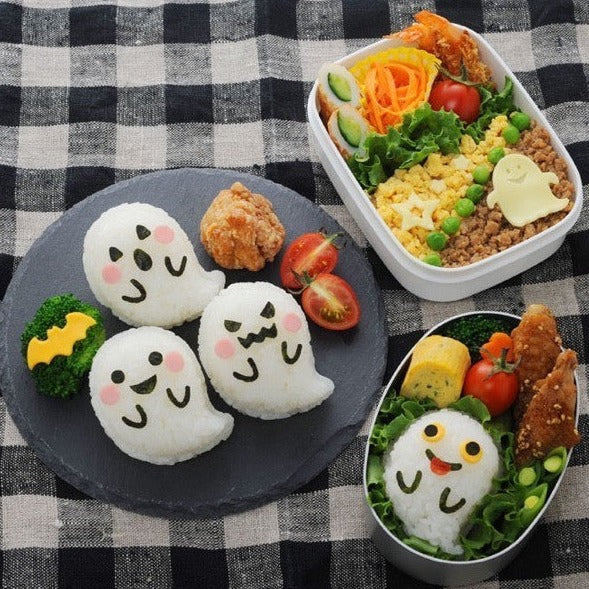 Four ghost-shaped rice balls with different nori expressions on a slate plate with fried chicken, cherry tomatoes, and broccoli, alongside two bento boxes with ghost onigiri and colorful side dishes, on a black gingham cloth