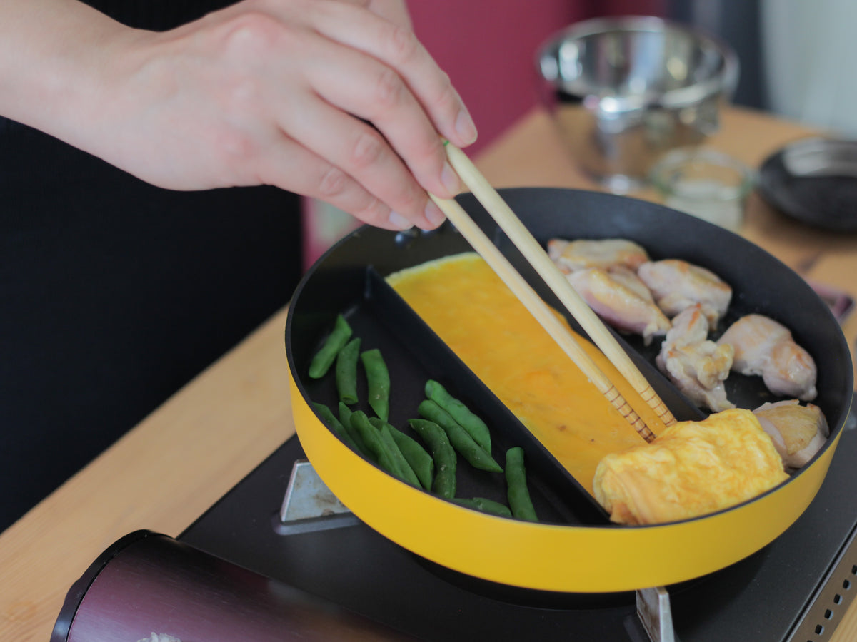 Hands using chopsticks to cook in Triple Pan yellow divided frying pan – tamagoyaki rolling in center, chicken wings on right, green beans on left, on portable induction hob