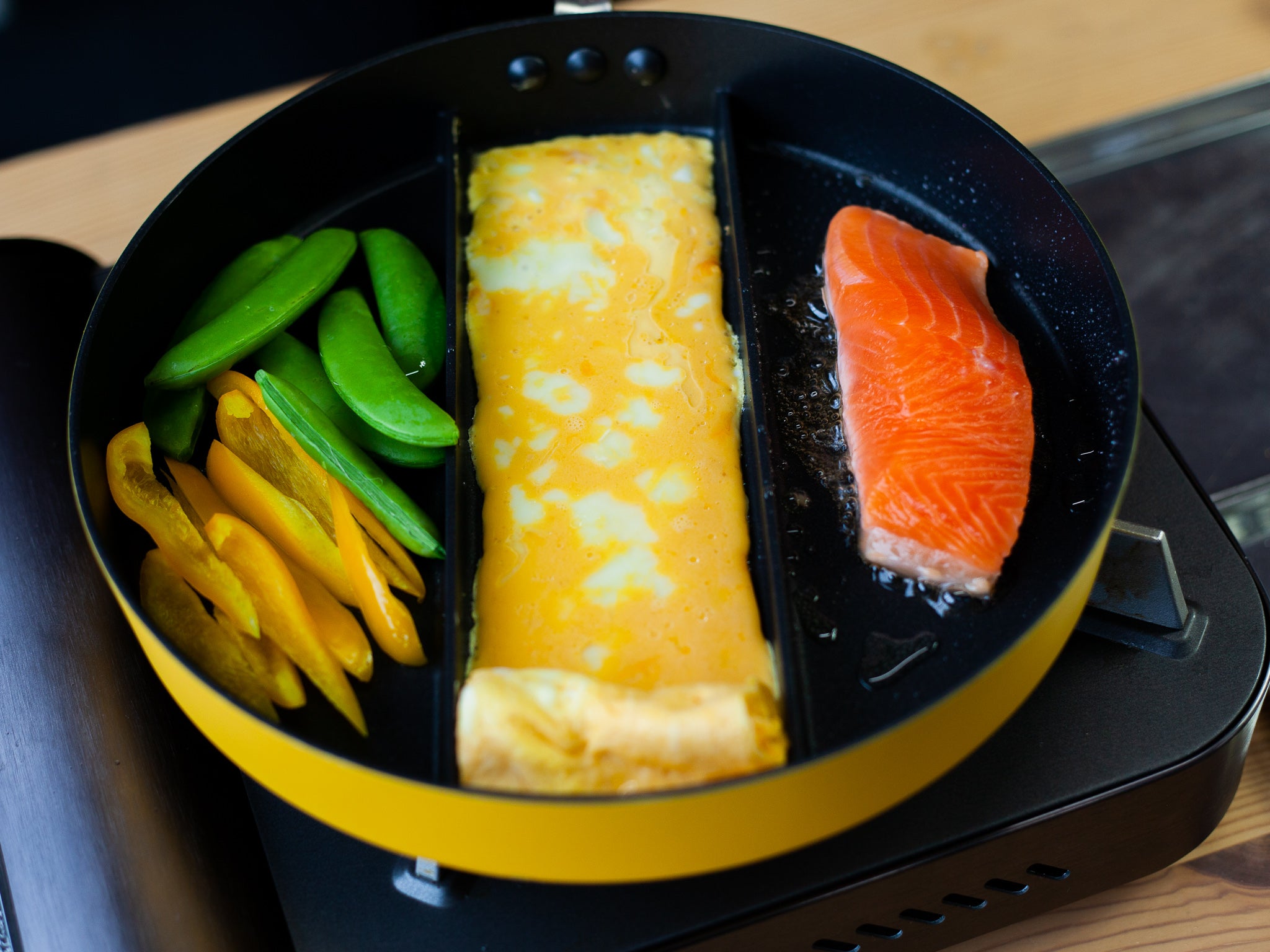 Triple Pan yellow divided frying pan with completed bento ingredients – rolled tamagoyaki in center section, salmon fillet on right, snap peas and yellow bell pepper on left, on portable induction hob