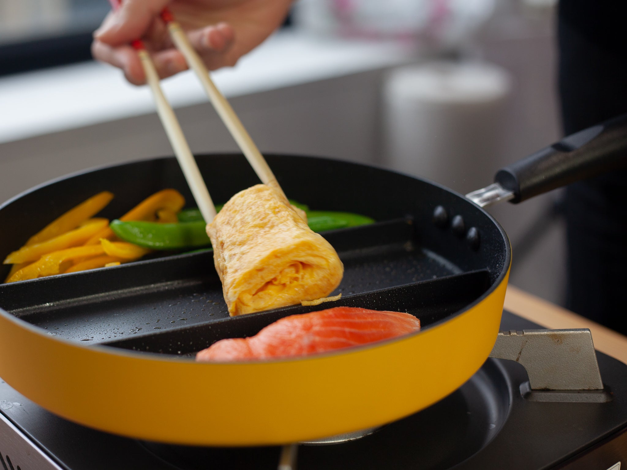 Close-up of Triple Pan yellow divided frying pan – hand lifting rolled tamagoyaki with chopsticks while snap peas, yellow bell pepper, and salmon cook in adjacent sections on induction hob