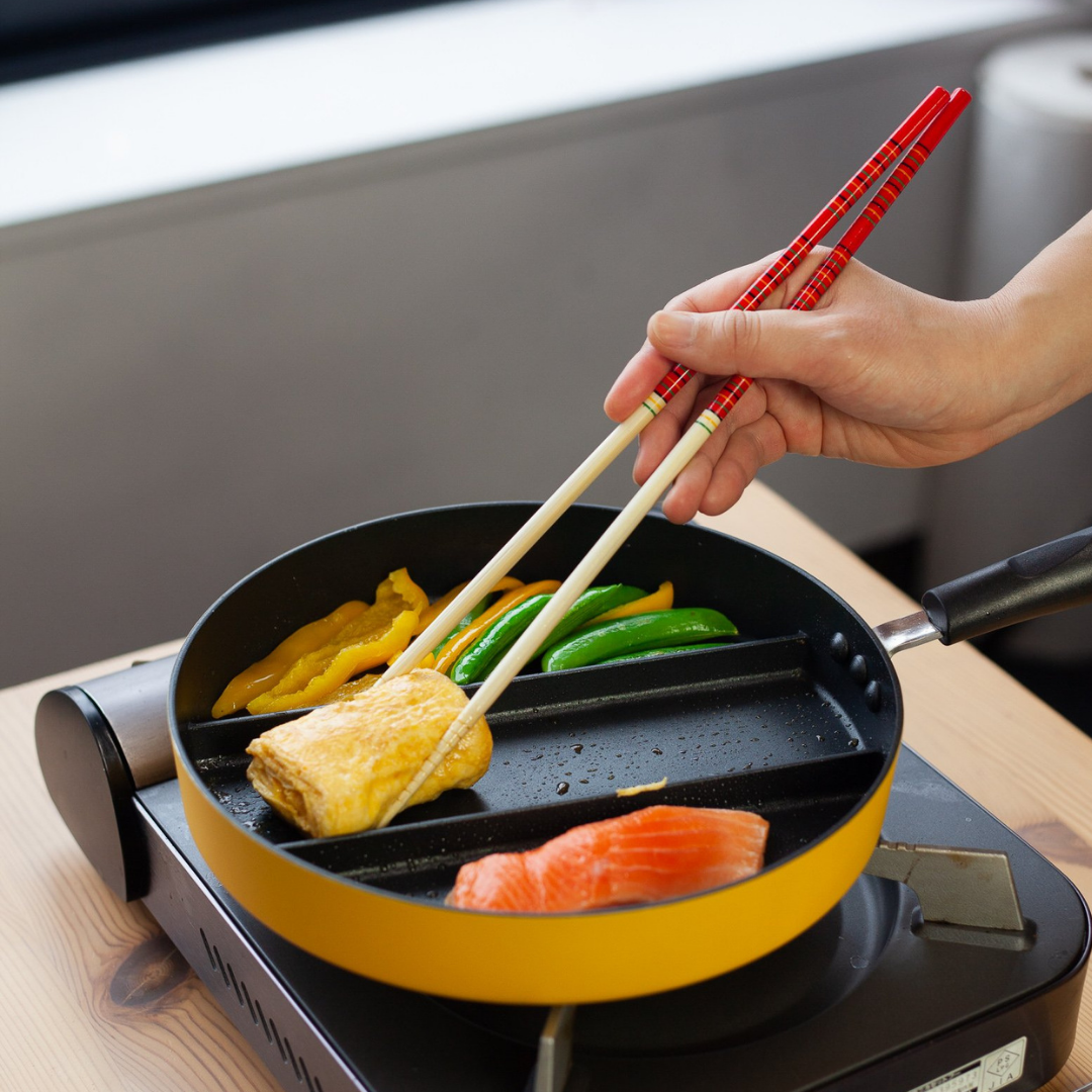 Triple Pan yellow divided frying pan on induction hob – cooking tamagoyaki, snap peas, yellow bell pepper, and salmon fillet simultaneously in three sections, hand using red chopsticks
