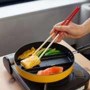 Triple Pan yellow divided frying pan on induction hob – cooking tamagoyaki, snap peas, yellow bell pepper, and salmon fillet simultaneously in three sections, hand using red chopsticks